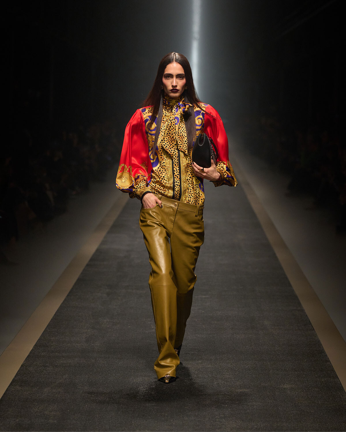 Model walks the runway in a vibrant red-and-gold patterned blouse with tan leather pants, carrying a small black clutch.