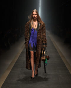 Model walking a fashion runway in a leopard-print coat over a purple dress, carrying a metallic bag under spotlight