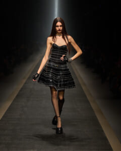 Model walking a runway in a black strapless sequined dress with black gloves and a silver necklace.