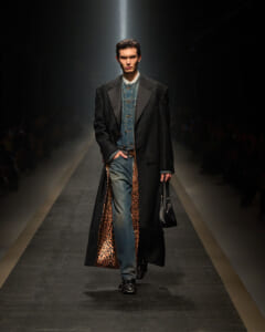 Male model walks a fashion runway wearing a long black patterned coat with leopard-print lining, a denim shirt, blue jeans, and a black bag.