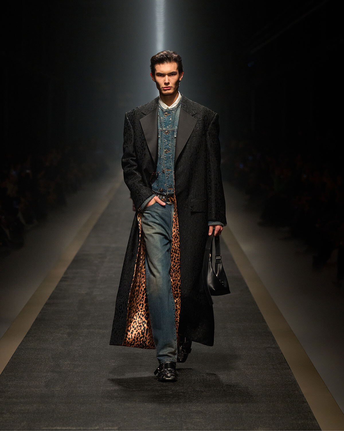 Male model walks a fashion runway wearing a long black patterned coat with leopard-print lining, a denim shirt, blue jeans, and a black bag.