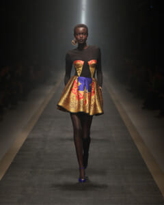 Model on a runway wearing a black bodice with a vibrant gold-and-red corset and a voluminous blue-gold patterned skirt, with large circular earrings.