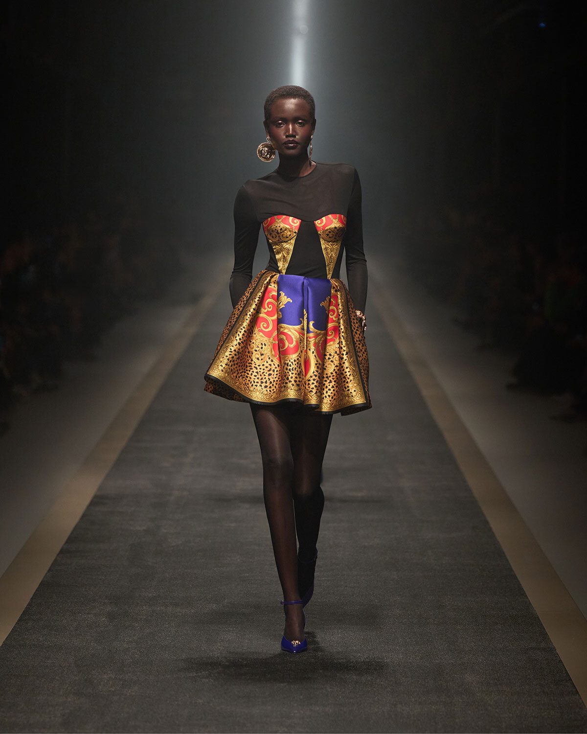 Model on a runway wearing a black bodice with a vibrant gold-and-red corset and a voluminous blue-gold patterned skirt, with large circular earrings.
