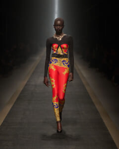 Model walks a fashion runway wearing a red, gold, and blue patterned bodysuit with black sheer sleeves and a chunky gold necklace.