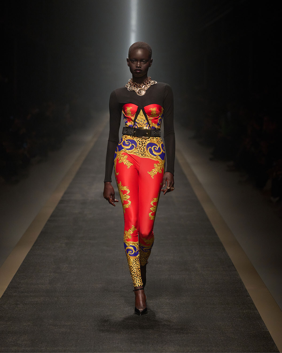 Model walks a fashion runway wearing a red, gold, and blue patterned bodysuit with black sheer sleeves and a chunky gold necklace.
