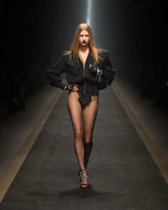 Model walking a fashion runway in an oversized black zip-front sweater, sheer black stockings, and strappy heels under a spotlight.