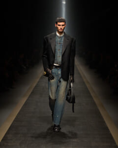 Male model walks a fashion runway in a dark, textured blazer over a denim jacket and jeans, hands gloved, carrying a black bag under spotlight.