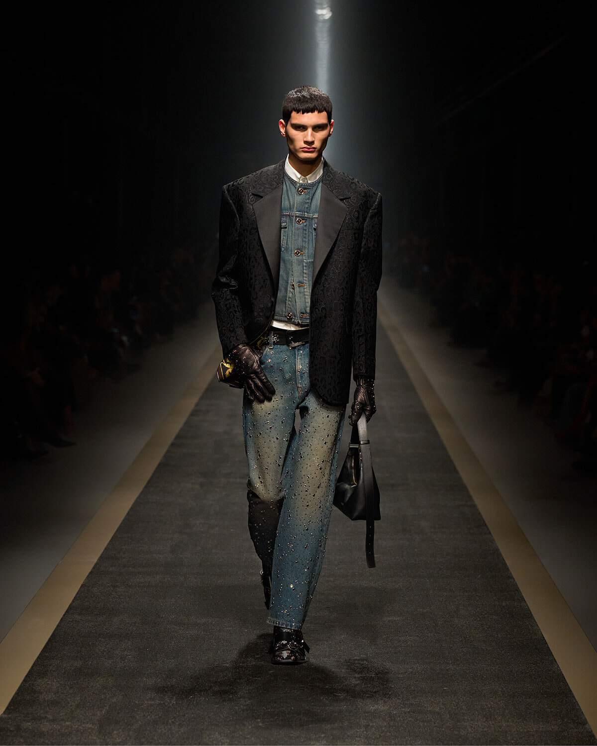 Male model walks a fashion runway in a dark, textured blazer over a denim jacket and jeans, hands gloved, carrying a black bag under spotlight.