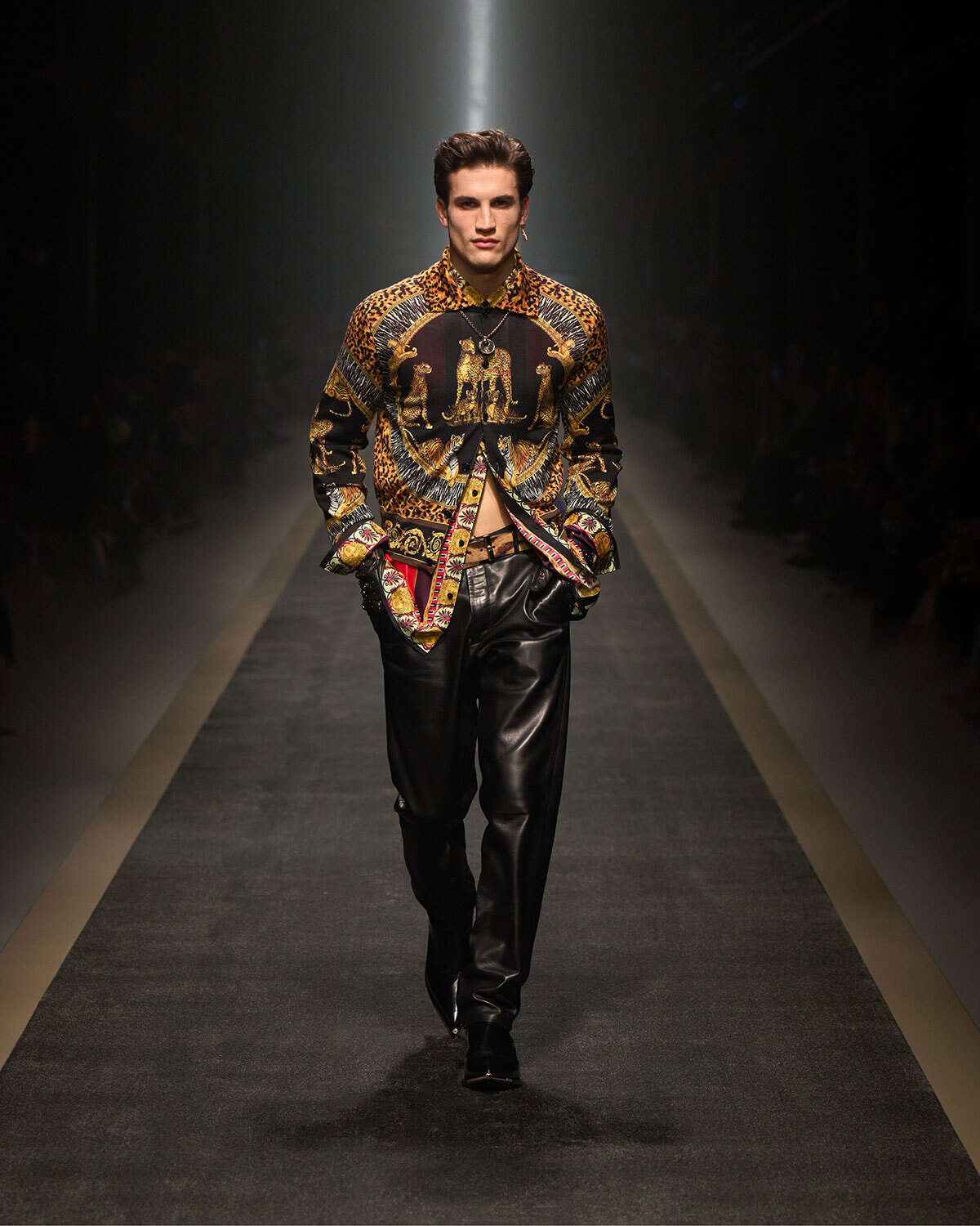 Male model on a runway wearing a bold patterned shirt with leopard print and gold motifs, paired with black leather pants.
