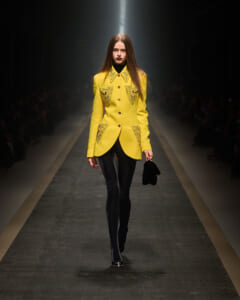 Model walks the runway in a bright yellow tailored coat with embroidered pockets, paired with black tights and a small black handbag under spotlight.
