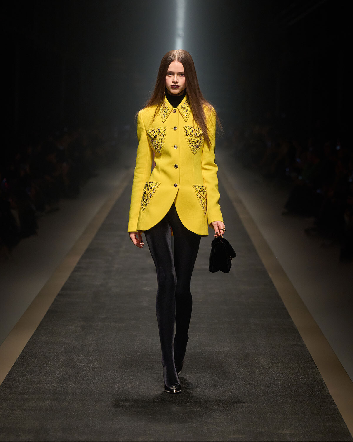 Model walks the runway in a bright yellow tailored coat with embroidered pockets, paired with black tights and a small black handbag under spotlight.