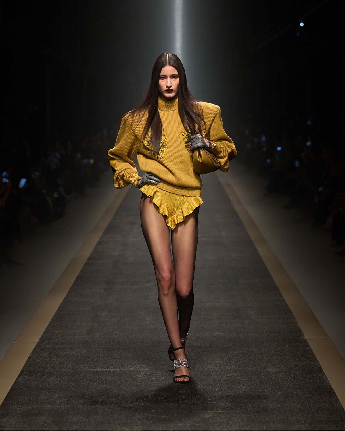 Model walks a fashion runway in a mustard oversized sweater and bright yellow ruffled shorts, with sheer tights and glittery sandals, carrying a tan bag under spotlight.