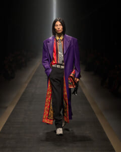 Model walking a fashion runway in a long purple coat with red-gold patterned lining, black trousers, and snakeskin shoes under spotlight.