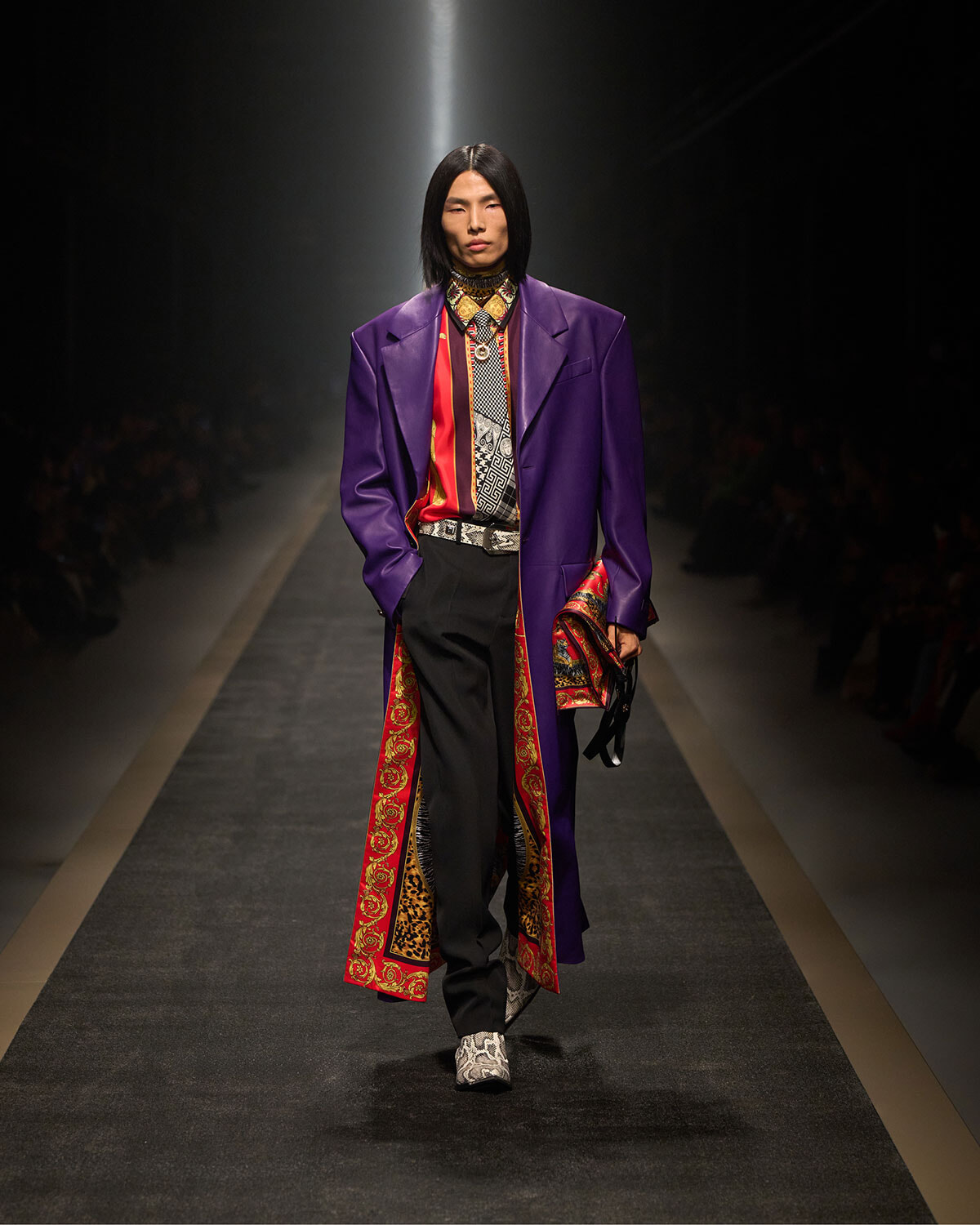 Model walking a fashion runway in a long purple coat with red-gold patterned lining, black trousers, and snakeskin shoes under spotlight.