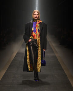 Model walking a runway in a long black coat, purple top, leopard scarf, and black leather pants with blue shoes and a blue bagraillet.