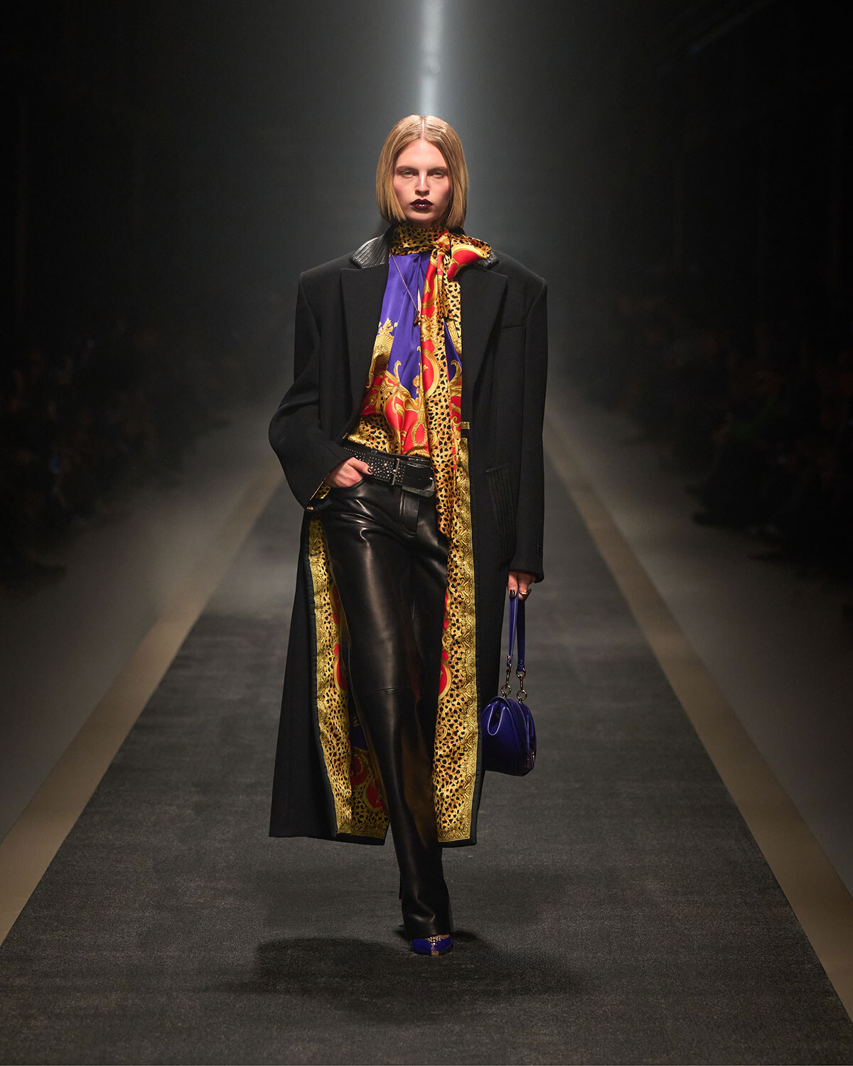 Model walking a runway in a long black coat, purple top, leopard scarf, and black leather pants with blue shoes and a blue bagraillet.
