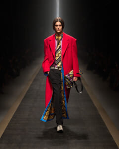 Model walking the runway in a bright red oversized coat with a patterned scarf, black trousers, and ornate blue-gold trim at a fashion show.