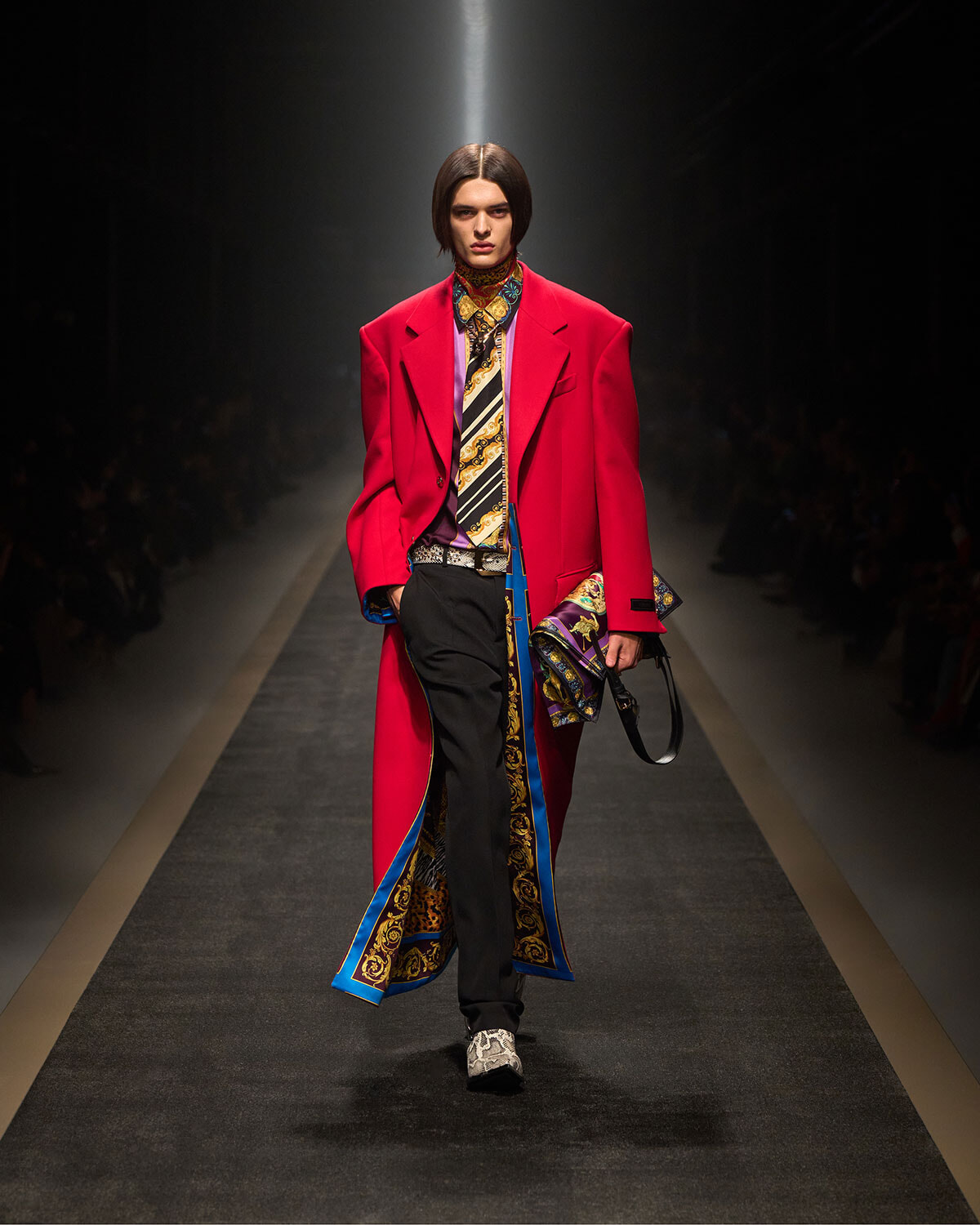 Model walking the runway in a bright red oversized coat with a patterned scarf, black trousers, and ornate blue-gold trim at a fashion show.