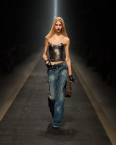 Model walks a fashion runway wearing a metallic strapless corset top, distressed blue jeans, black gloves, and a small leopard-print handbag.