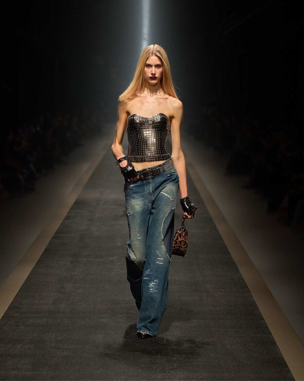 Model walks a fashion runway wearing a metallic strapless corset top, distressed blue jeans, black gloves, and a small leopard-print handbag.