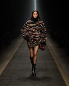 Model walking a runway in a chunky multicolor knit sweater with a fringed mini skirt, black boots, and a burgundy handbag in hand.