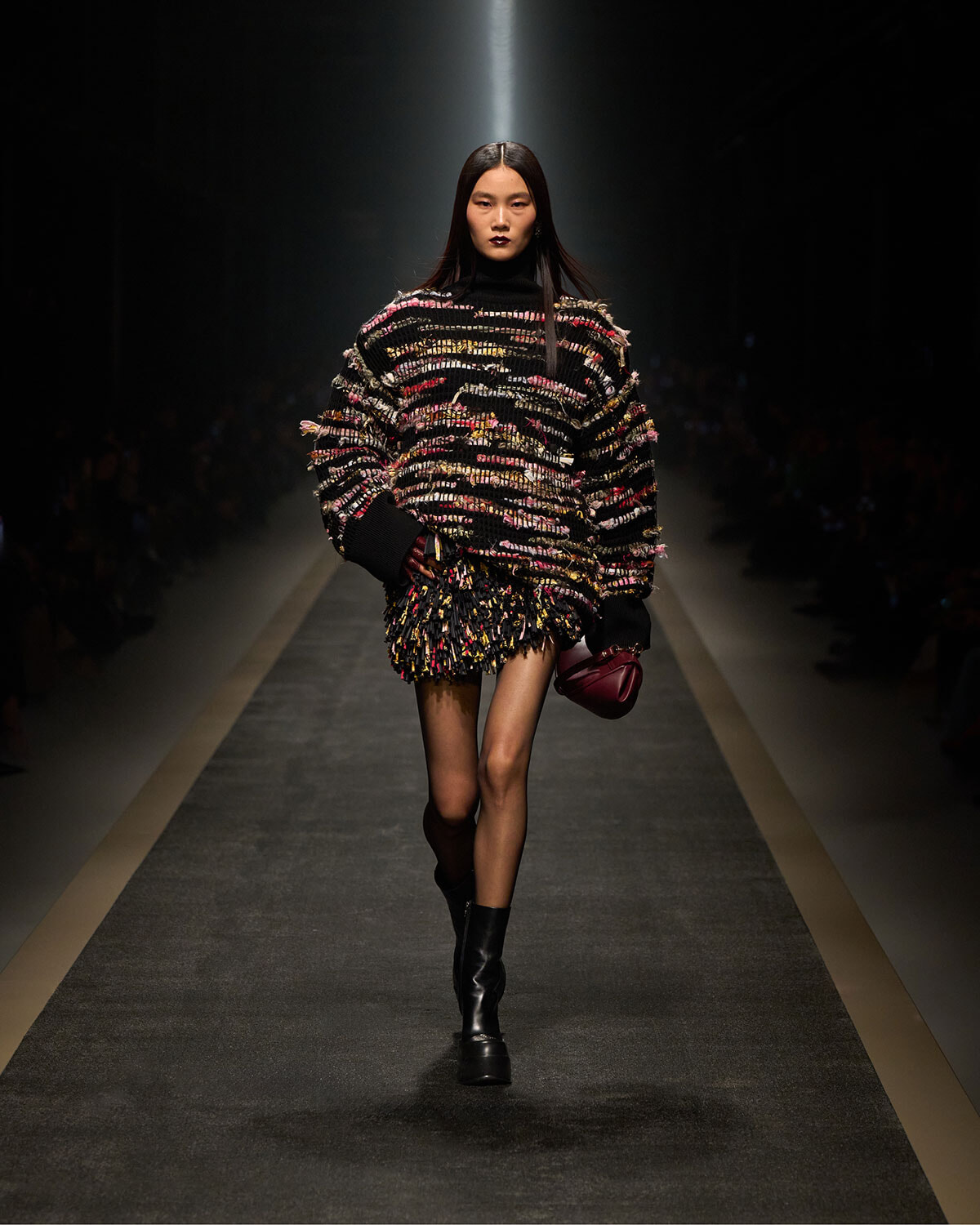 Model walking a runway in a chunky multicolor knit sweater with a fringed mini skirt, black boots, and a burgundy handbag in hand.
