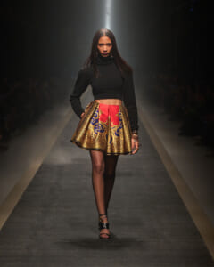 Model on a runway wearing a black cropped turtleneck and a gold-and-red patterned mini skirt. - Spotted/walks the catwalk in a sleek black top and a vibrant printed gold skirt.