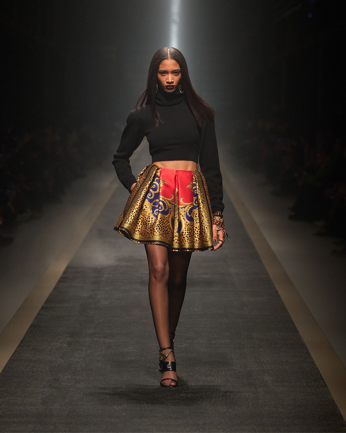 Model on a runway wearing a black cropped turtleneck and a gold-and-red patterned mini skirt. - Spotted/walks the catwalk in a sleek black top and a vibrant printed gold skirt.