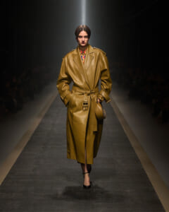 Model walks the runway in a long tan leather trench coat with a belt, spotlight on the catwalk.