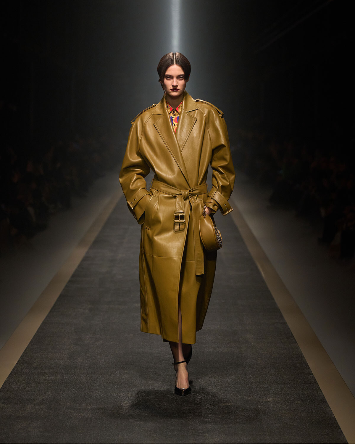 Model walks the runway in a long tan leather trench coat with a belt, spotlight on the catwalk.