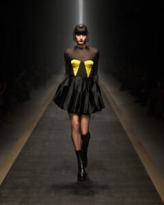 Model walks the runway in a black sheer top with a yellow, corset-style bodice and a voluminous black satin skirt, knee-high boots.