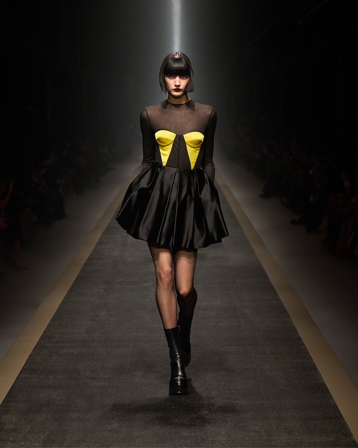Model walks the runway in a black sheer top with a yellow, corset-style bodice and a voluminous black satin skirt, knee-high boots.