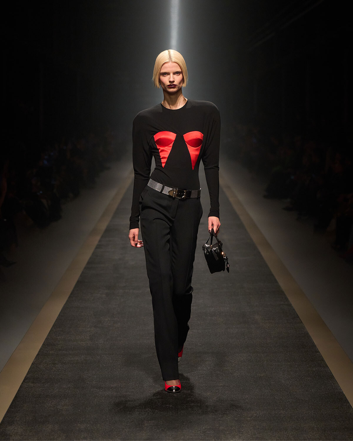 Model on runway wearing a black long-sleeve top with red chest cutouts, black trousers, a metallic belt, red heels, and a small black handbag.