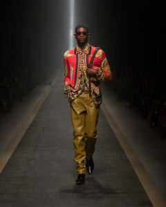 Male model on a runway wearing a bold red patterned shirt, gold leather pants, sunglasses, and a shoulder strap bag.
