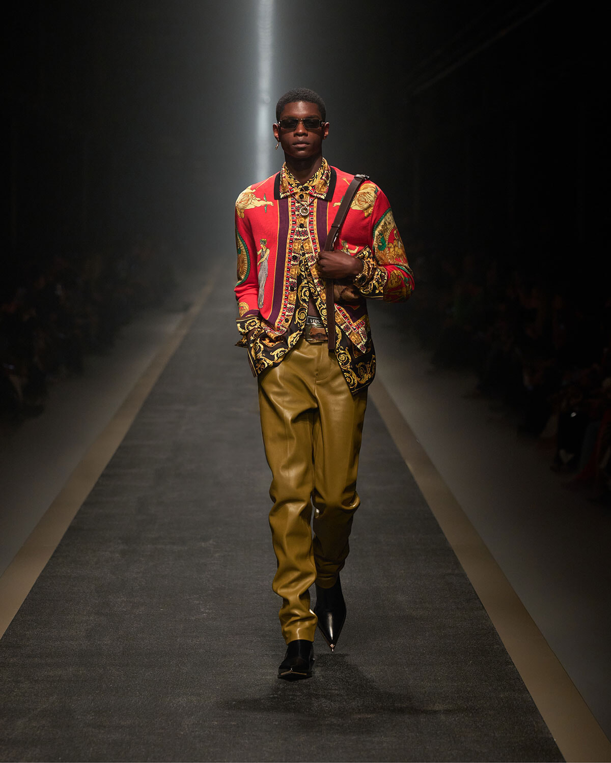 Male model on a runway wearing a bold red patterned shirt, gold leather pants, sunglasses, and a shoulder strap bag.