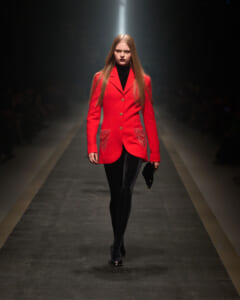 Model walking a runway in a striking red tailored coat with gold buttons and heart-embellished pockets, black turtleneck and tights.