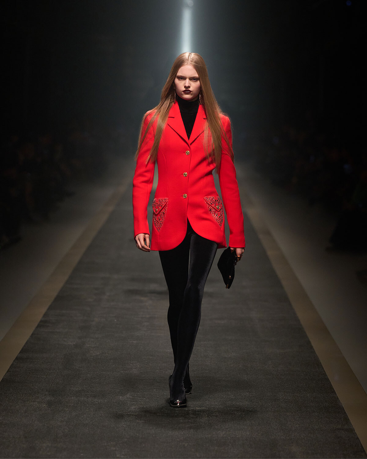 Model walking a runway in a striking red tailored coat with gold buttons and heart-embellished pockets, black turtleneck and tights.