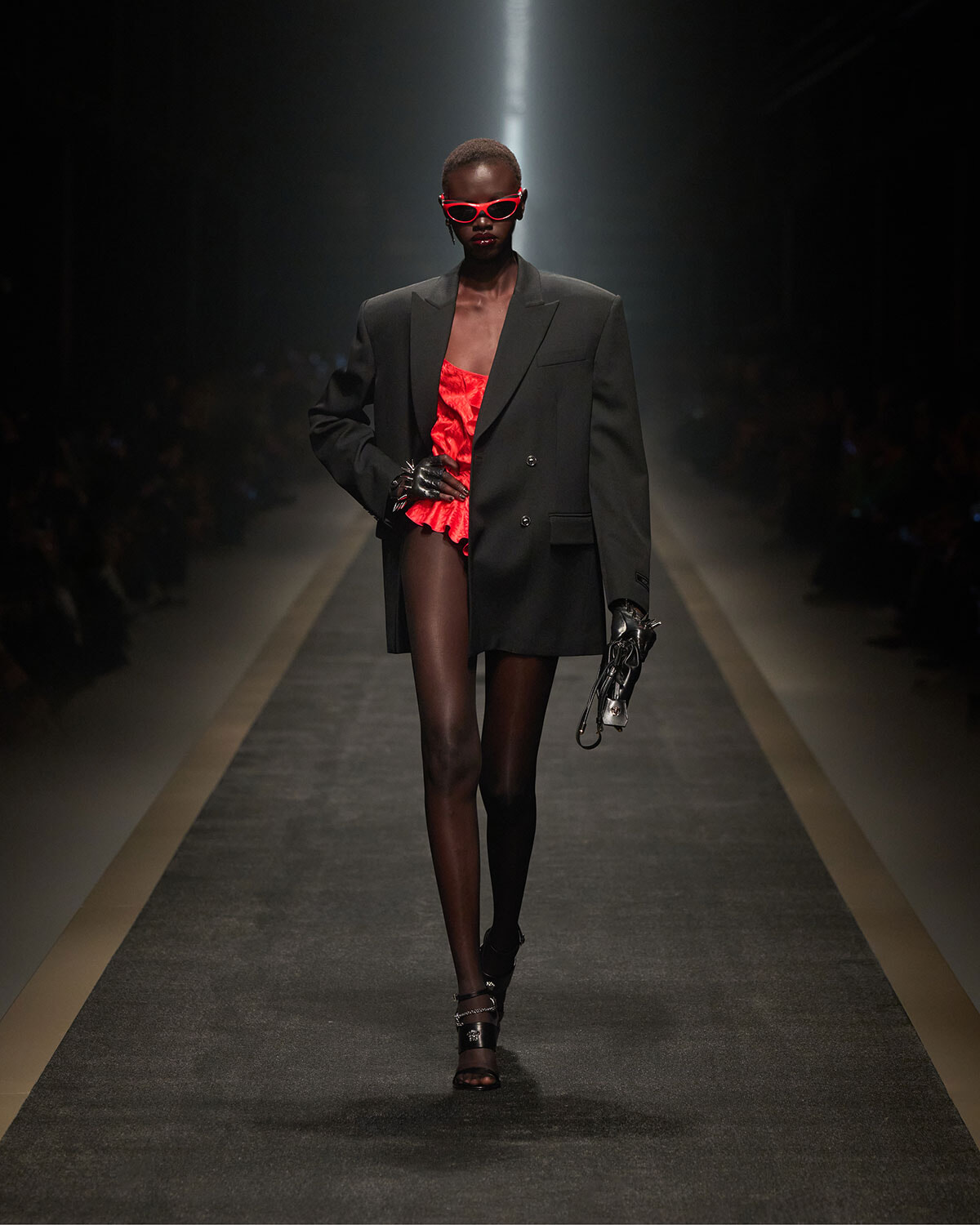 Model walks the runway in an oversized black blazer over a red ruffled bodysuit, red sunglasses, and black strappy heels under a spotlight.