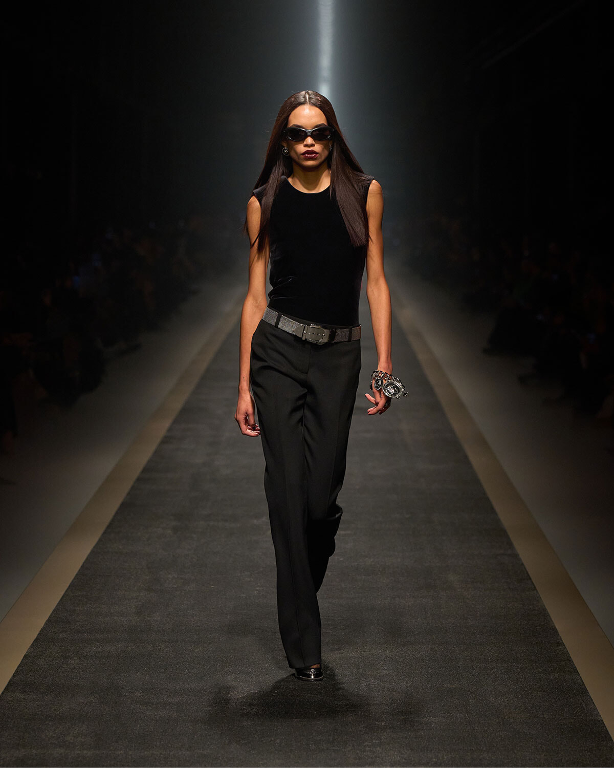 Model walks a runway in a black sleeveless top, black trousers, a gray belt, dark sunglasses, and a chunky bracelet under spotlight.