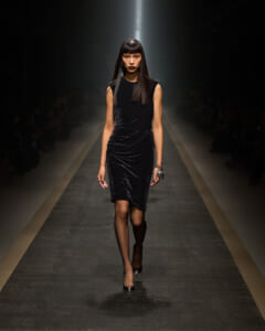 Female model walking a runway in a sleeveless black velvet dress with draped folds, spotlight behind her, wearing black heels and bracelets.
