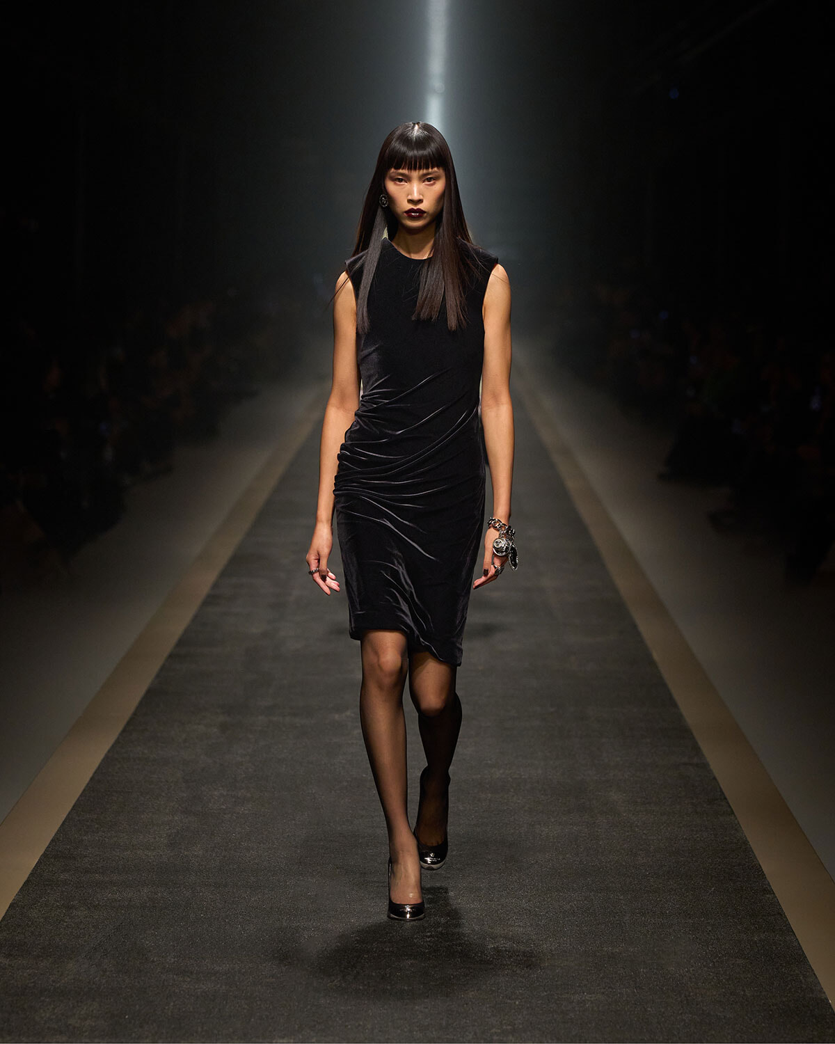Female model walking a runway in a sleeveless black velvet dress with draped folds, spotlight behind her, wearing black heels and bracelets.