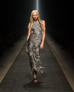 Female model walks the runway in a shimmering silver textured gown with fringe at the hem and a spotlight overhead.