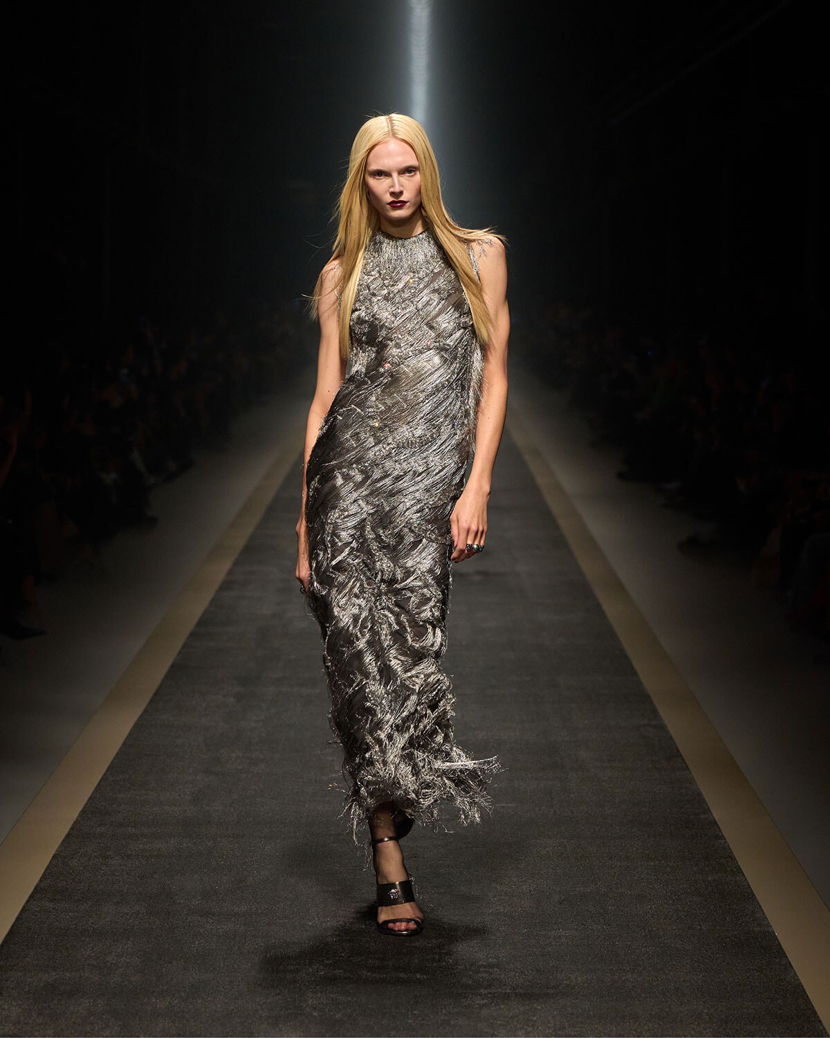 Female model walks the runway in a shimmering silver textured gown with fringe at the hem and a spotlight overhead.