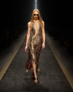 Model walks a metallic gold fringe dress down a runway, wearing dark sunglasses and gold heels under spotlight.