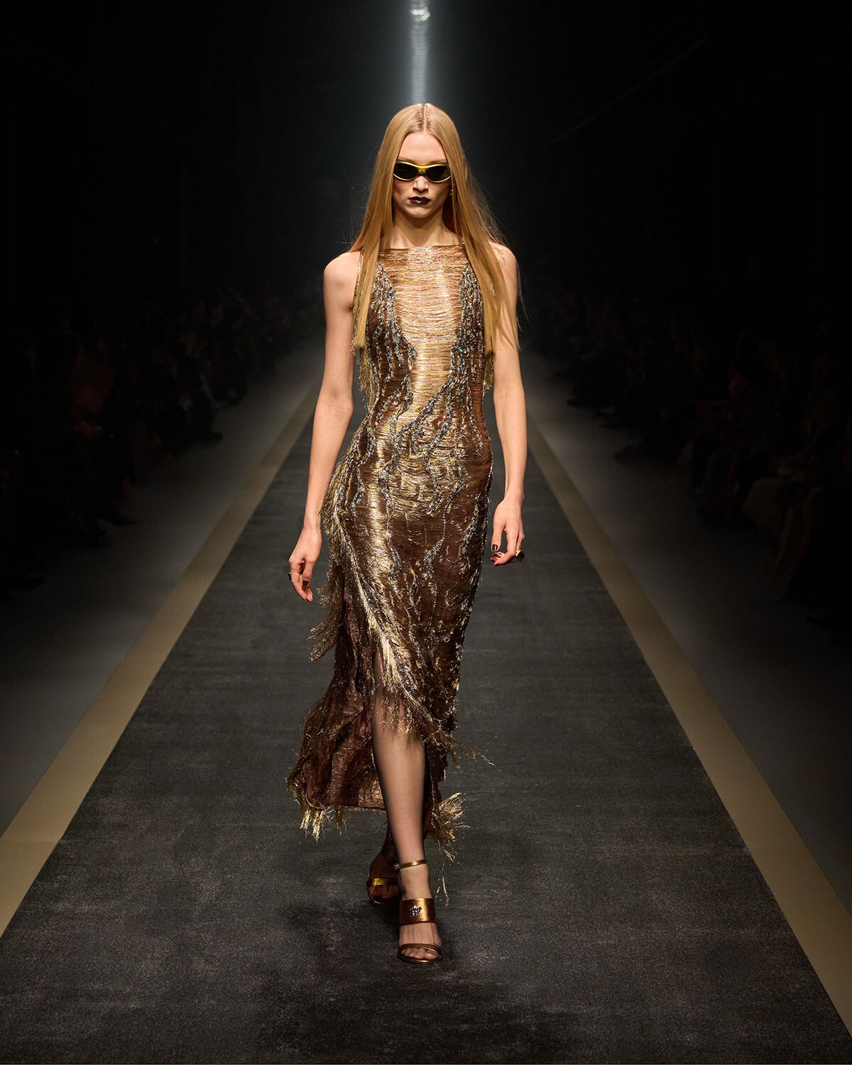 Model walks a metallic gold fringe dress down a runway, wearing dark sunglasses and gold heels under spotlight.