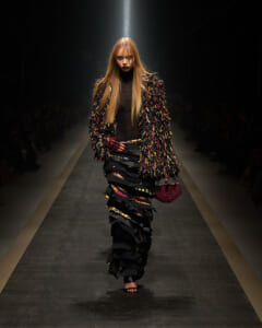 Model walks a fashion runway wearing a black fringed, sequined coat with multicolor accents, paired with a black turtleneck and layered skirt; burgundy handbag in hand under a spotlight.