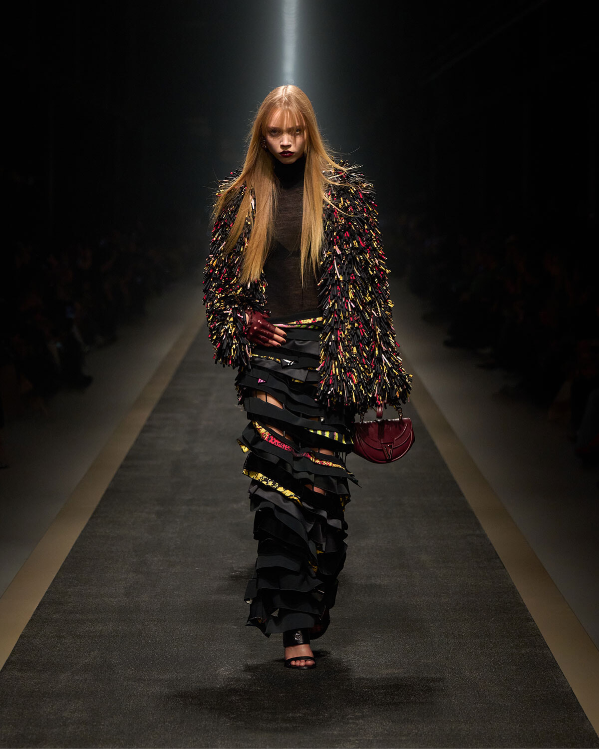 Model walks a fashion runway wearing a black fringed, sequined coat with multicolor accents, paired with a black turtleneck and layered skirt; burgundy handbag in hand under a spotlight.