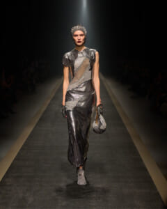 Model in a metallic silver gown walks a dim runway, wearing a spiked headpiece and matching accessories for a high-fashion show.