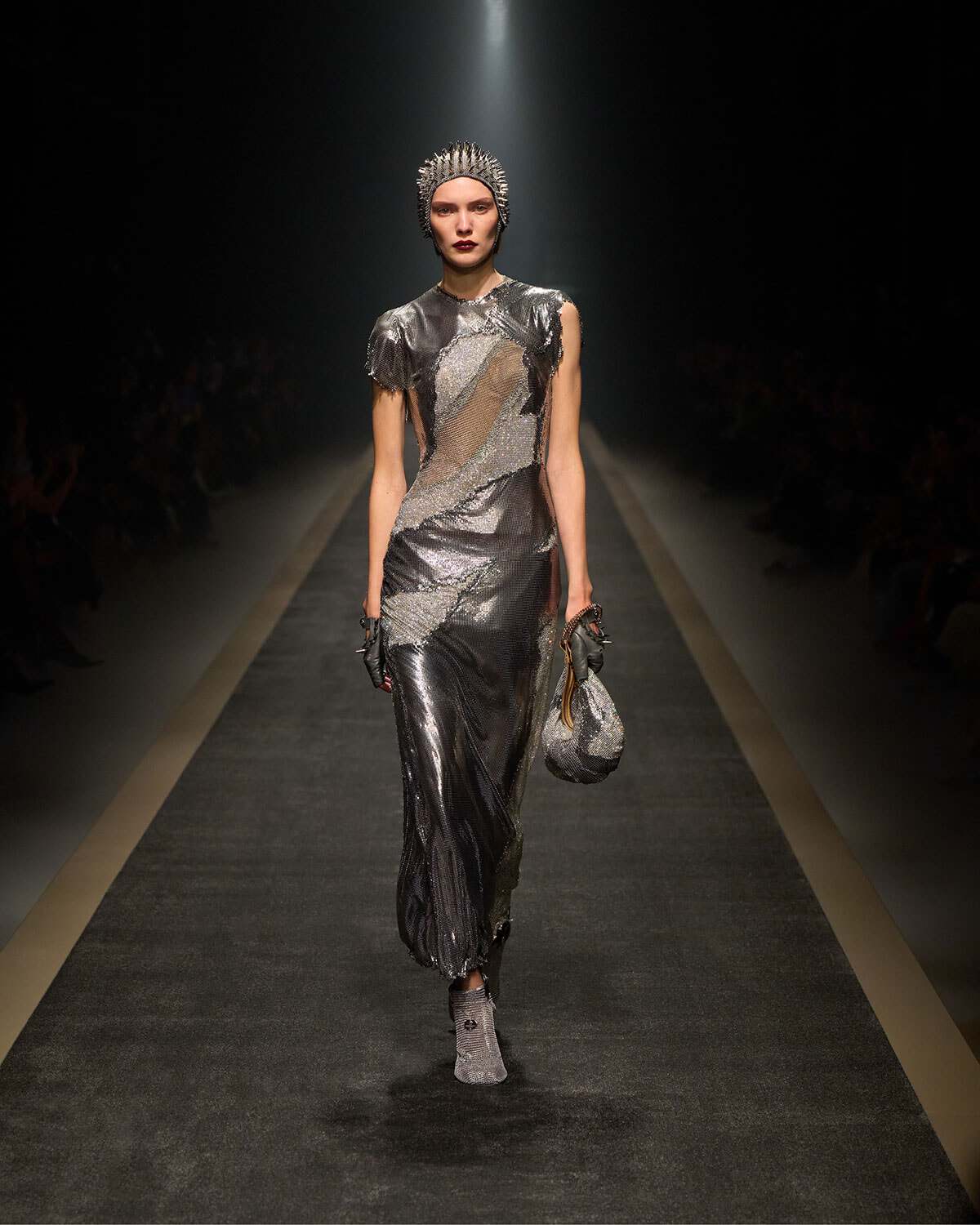 Model in a metallic silver gown walks a dim runway, wearing a spiked headpiece and matching accessories for a high-fashion show.