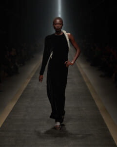 Model walks the runway in a floor-length black velvet dress with a jeweled strap over the shoulder on a spotlighted catwalk.